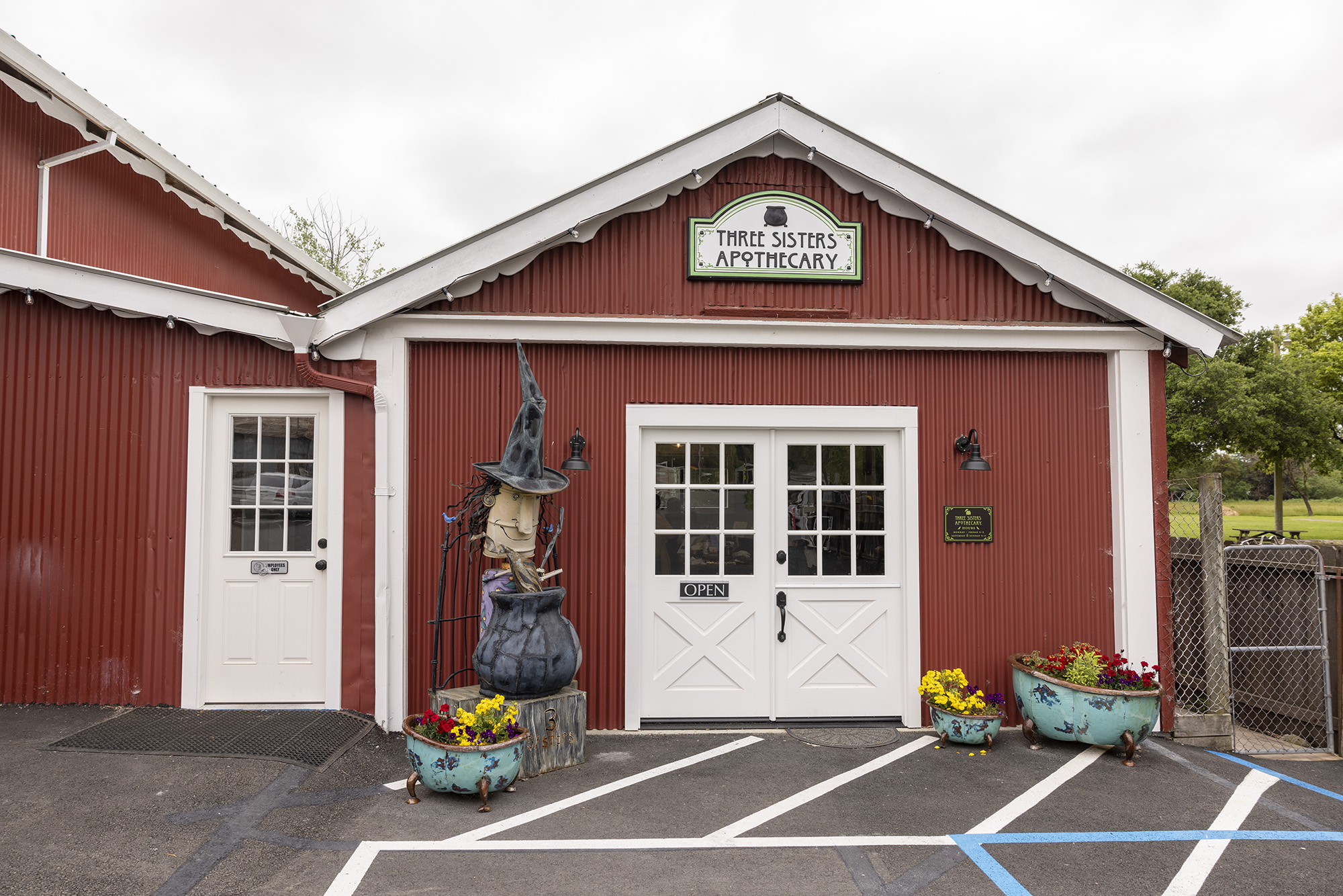 Three Sisters Apothecary exterior with sculpture of witch brewing over cauldron and bath tub shaped flowers pots with yellow, red, and purple flowers.