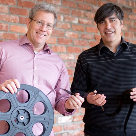 Two men holding film reel in front of brick wall