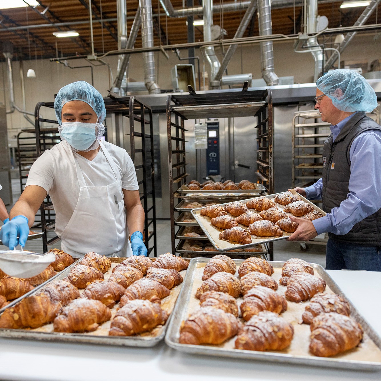 Baker applies confectioner sugar to freshly baked croissants while owner moves completed creations to racks.